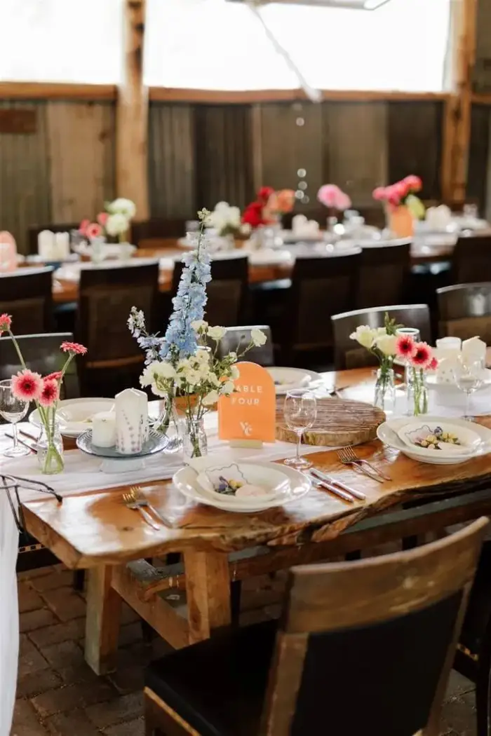 Reception table set with rustic timber furniture, gerbera daisies and delphinium in bud vases