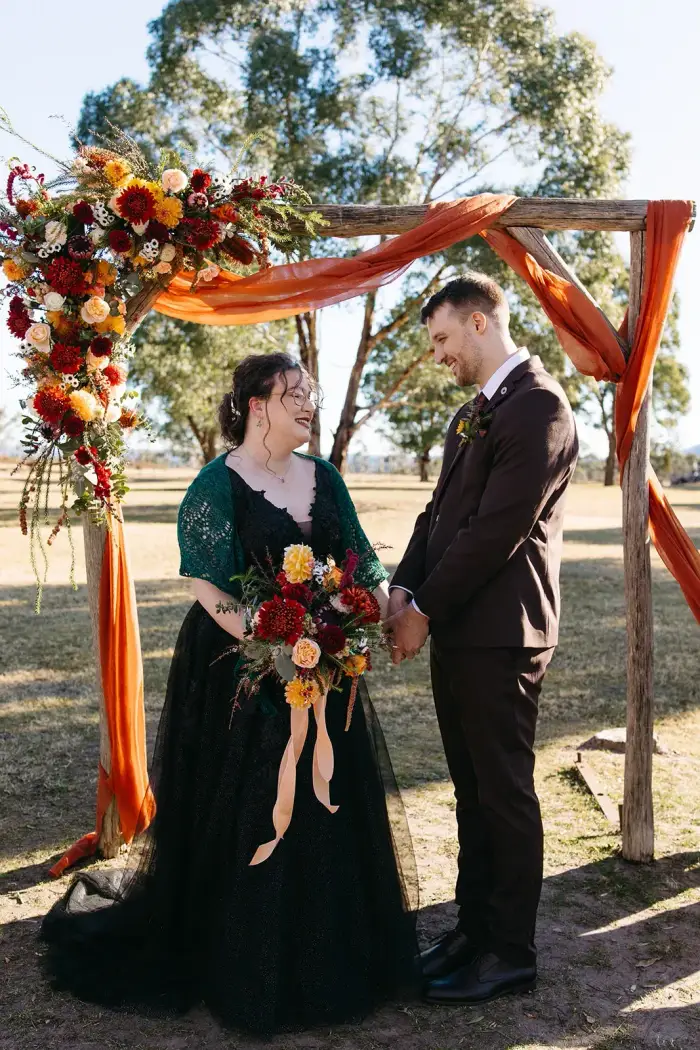 Bride and groom holding hands under an autumnal floral ceremony arch in deep red and burgundy tones